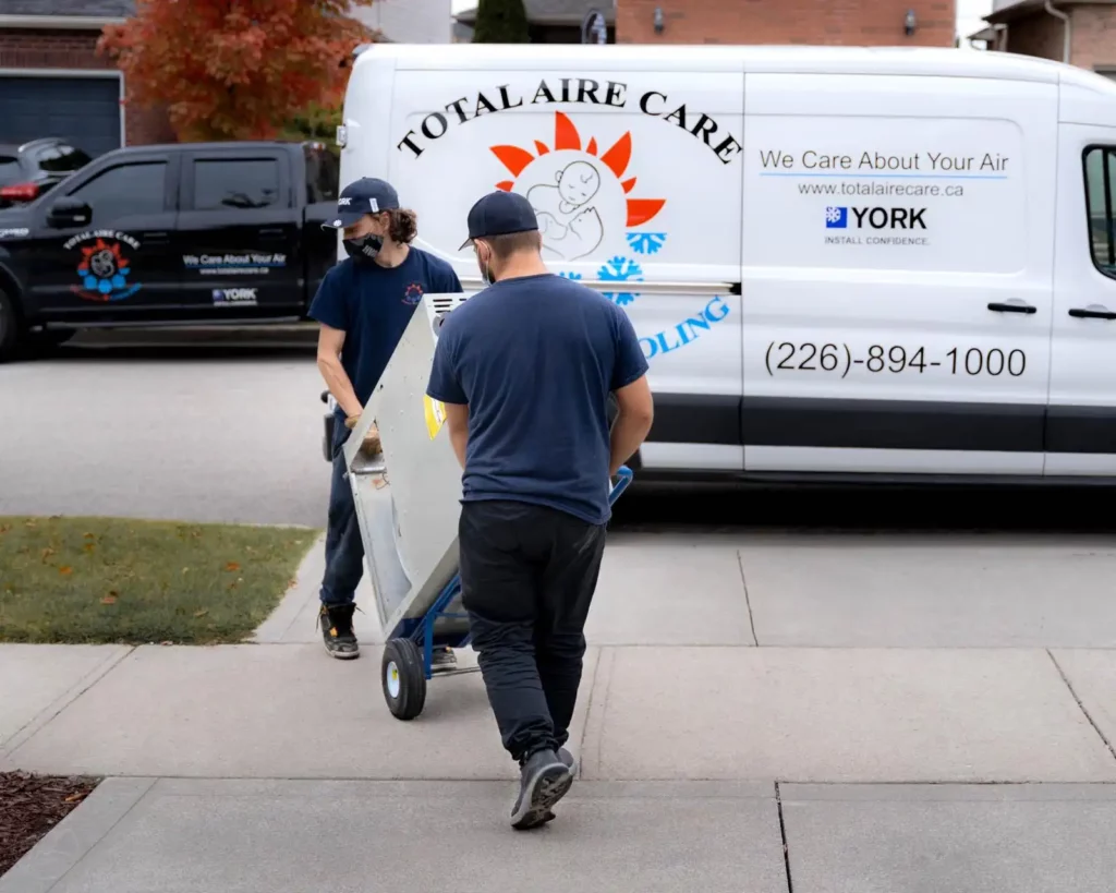 Two HVAC technicians from Total Aire Care, dressed in company uniforms, transport a new air conditioning unit on a dolly towards a residential home, with a branded service van displaying contact information parked in the driveway on an overcast day.