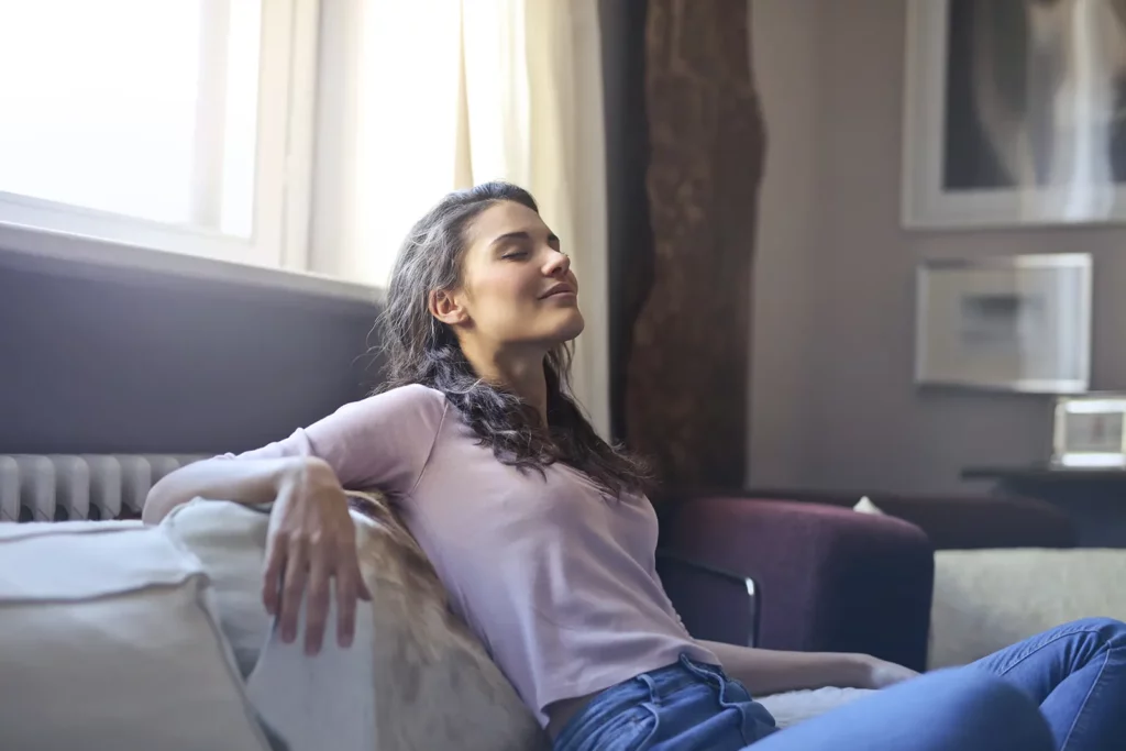 A woman relaxing on a couch with her eyes closed, enjoying the comfort of a well-ventilated room, possibly with a aprilaire humidifier running in the background.