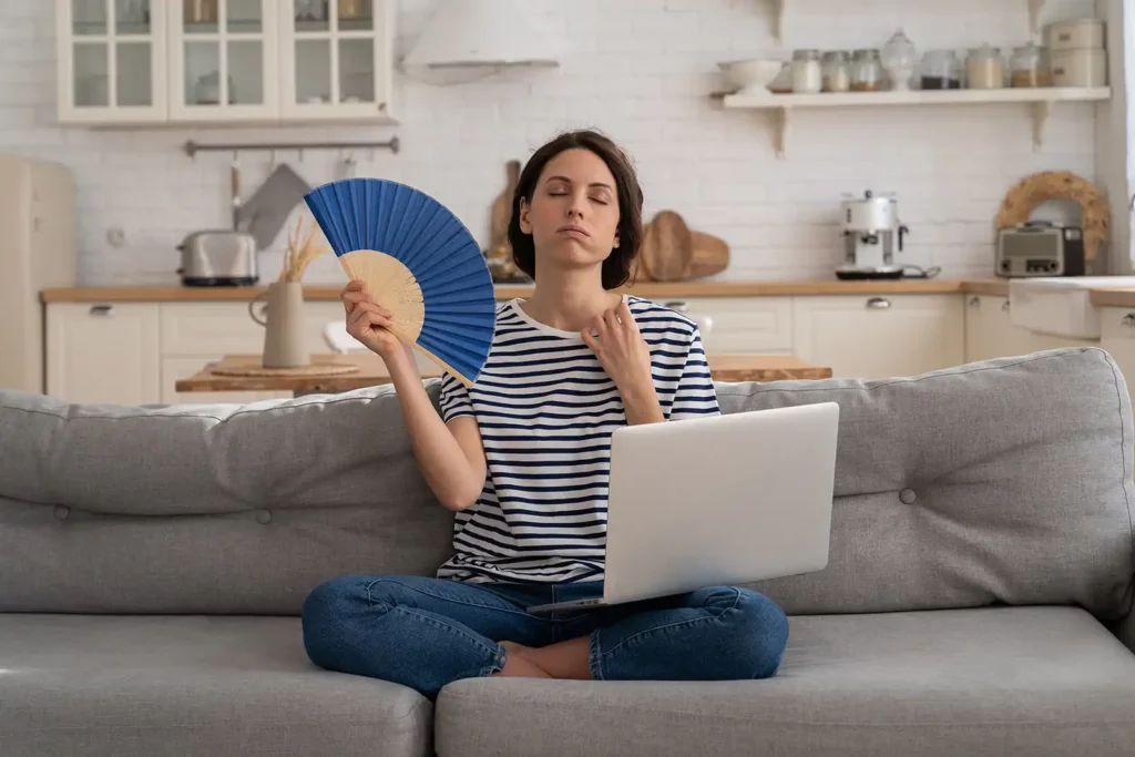 A woman sitting cross-legged on a gray sofa, fanning herself with a blue hand fan, and looking slightly distressed due to heat while using a laptop in a well-lit, modern kitchen setting. She is wearing a casual striped t-shirt and blue jeans, embodying a scene of discomfort likely due to a hot environment. This image may relate to topics like summer heat, air conditioning needs, or home comfort. Perfect for content addressing the importance of a well-functioning AC system during warm seasons.