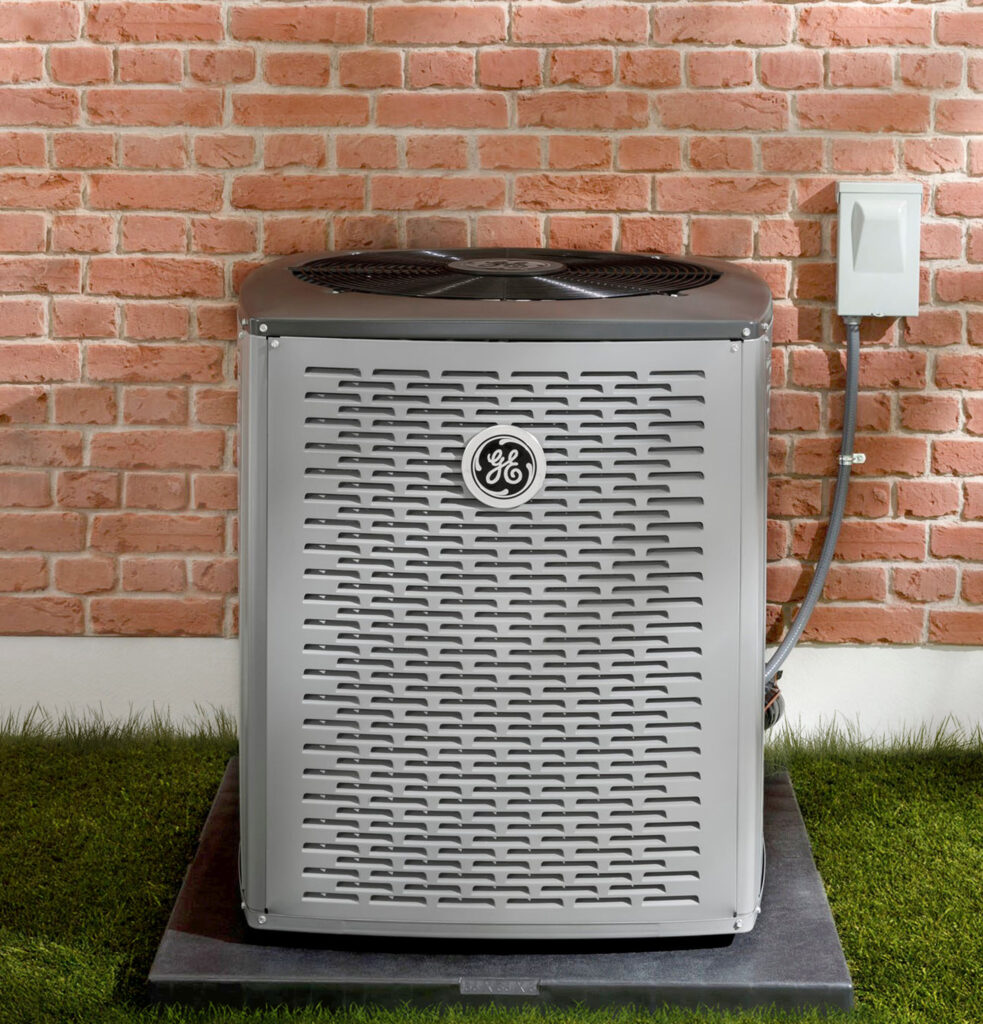 Image of a GE Heat Pump unit on a slab of concrete next to a red brick wall with fake grass.  