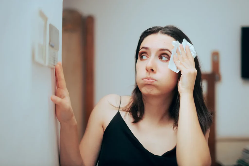 A young woman wiping sweat from her forehead with a tissue while looking up at an air conditioner thermostat in a room, appearing to be feeling hot and uncomfortable.