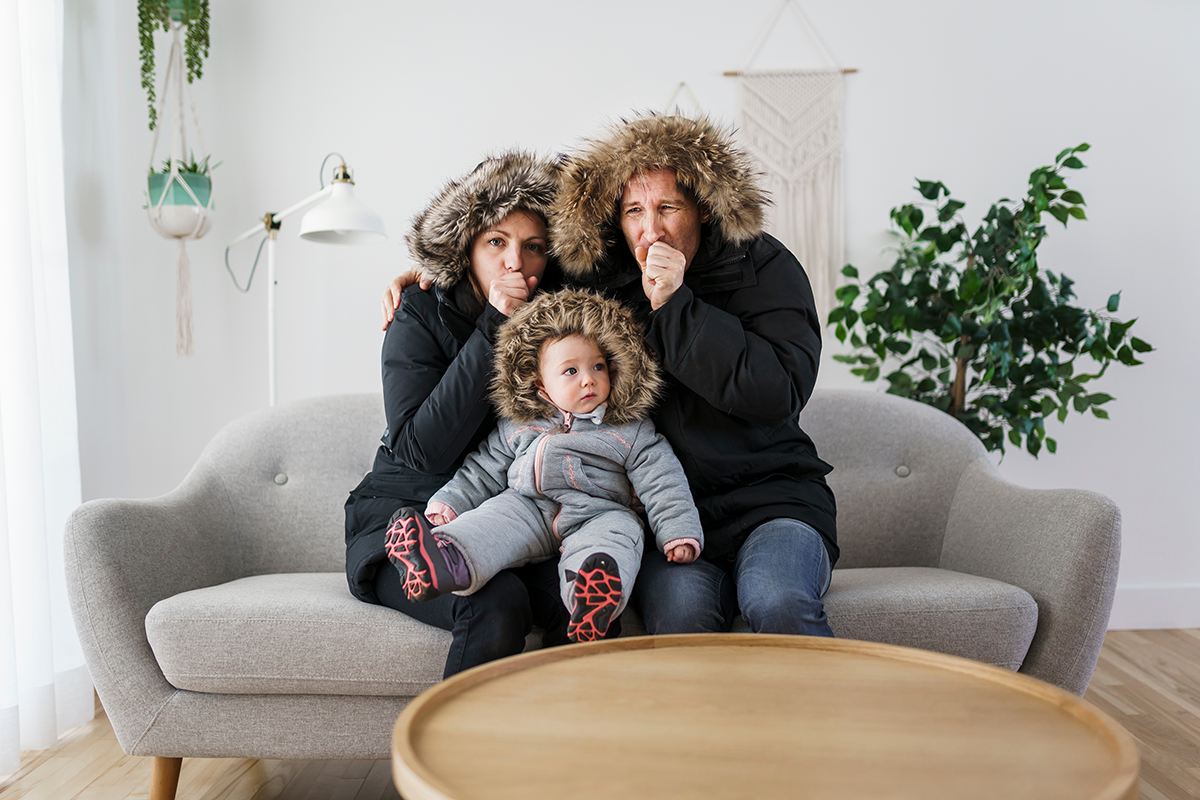 Family in winter coats sitting on a sofa indoors, mimicking being cold with one child in the center looking calm, surrounded by plants and minimalist decor, illustrating a cozy and quirky home setting