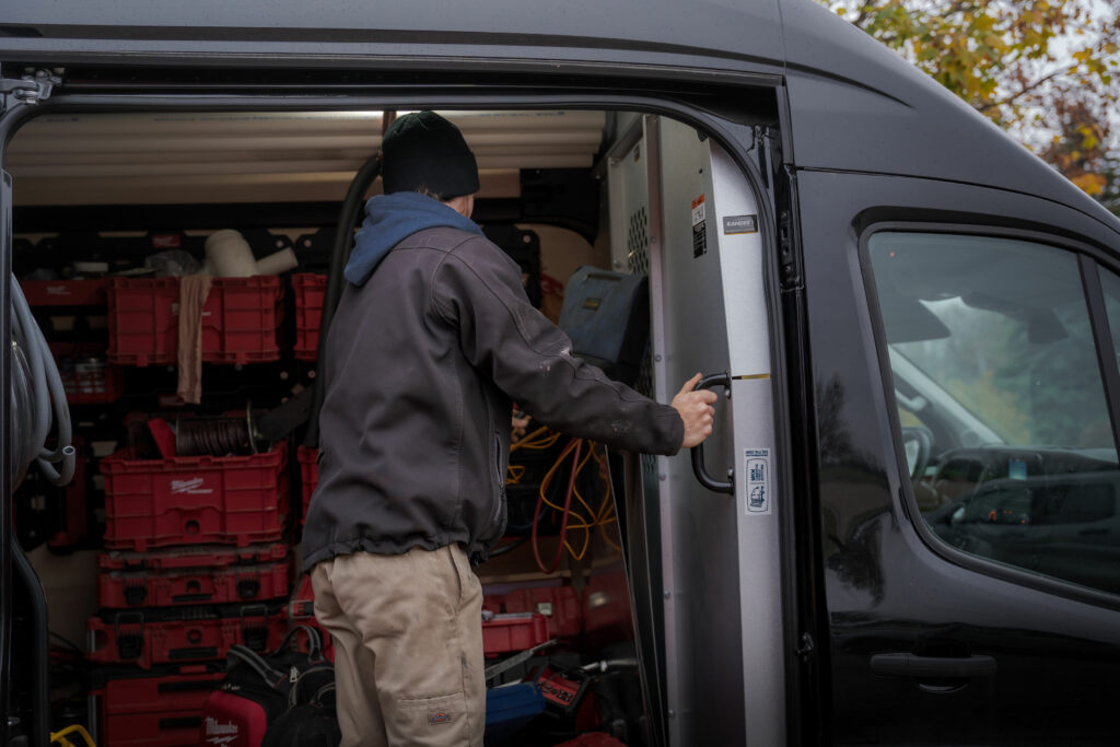A Total Aire Care employee, dressed in a grey jacket and beanie, is seen opening the side door of a well-organized work van filled with red toolboxes and various equipment. The image captures a moment of preparation, highlighting the organization and readiness of a mobile work environment. Regular maintenance is one energy saving tips.
