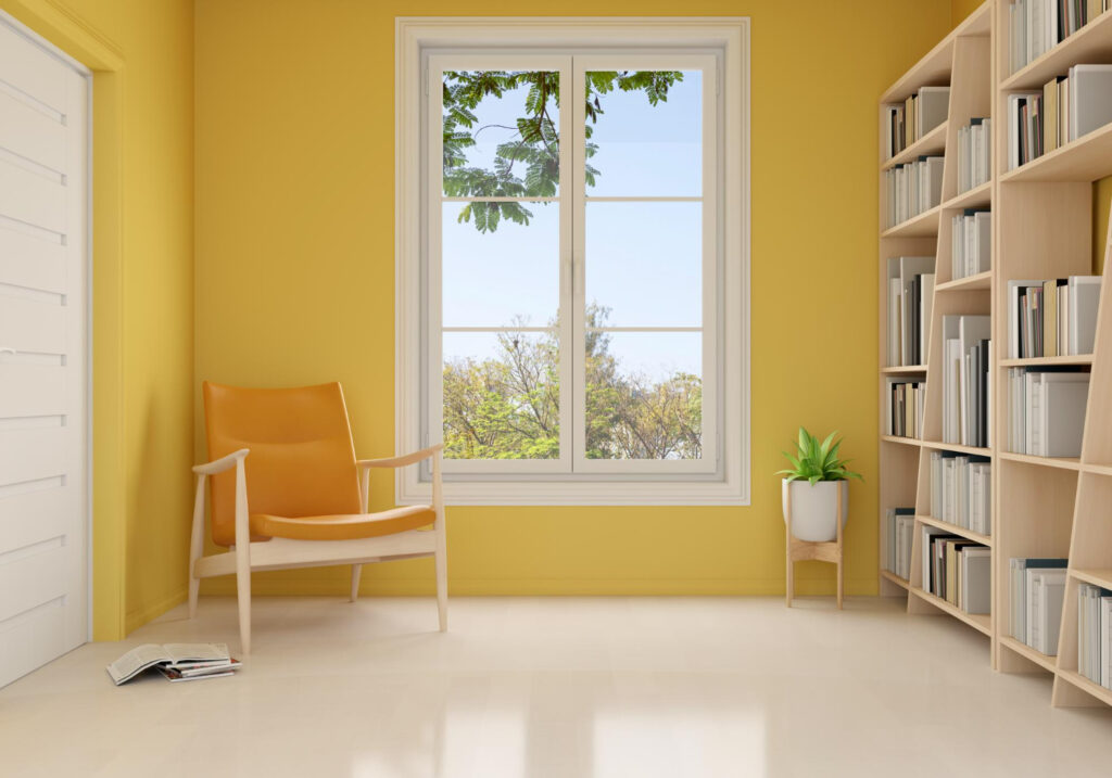 Bright and airy reading nook with a stylish orange armchair, a white bookshelf filled with books, and a large window with a view of green foliage, in a sunny yellow room.