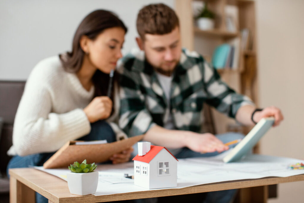 Medium shot of a focused young couple reviewing house plans indoors with a small model house and succulent plant on the table, investigating the Canada greener homes grant.