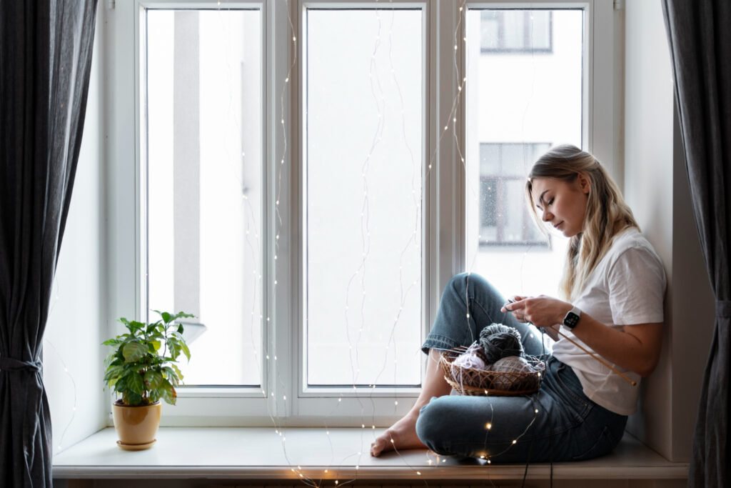 oung woman sitting by an energy saving window on a cozy windowsill, engaged in knitting from a basket, with a potted plant and fairy lights, promoting a relaxed hobby at home.