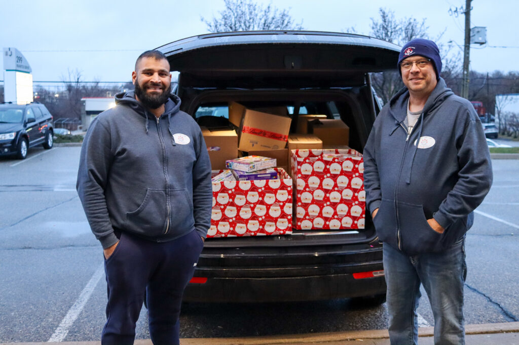 Two smiling men standing next to an open trunk of an SUV filled with boxes and gift-wrapped packages, likely participating in a charitable event or community assistance program.