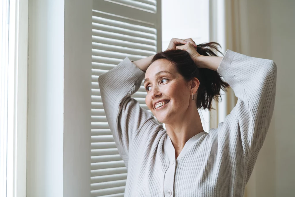 A joyful middle-aged woman, breathing clean air, with dark hair smiling and tying her hair up while standing in a room with natural light streaming through venetian blinds, conveying a sense of relaxation and happiness at home.