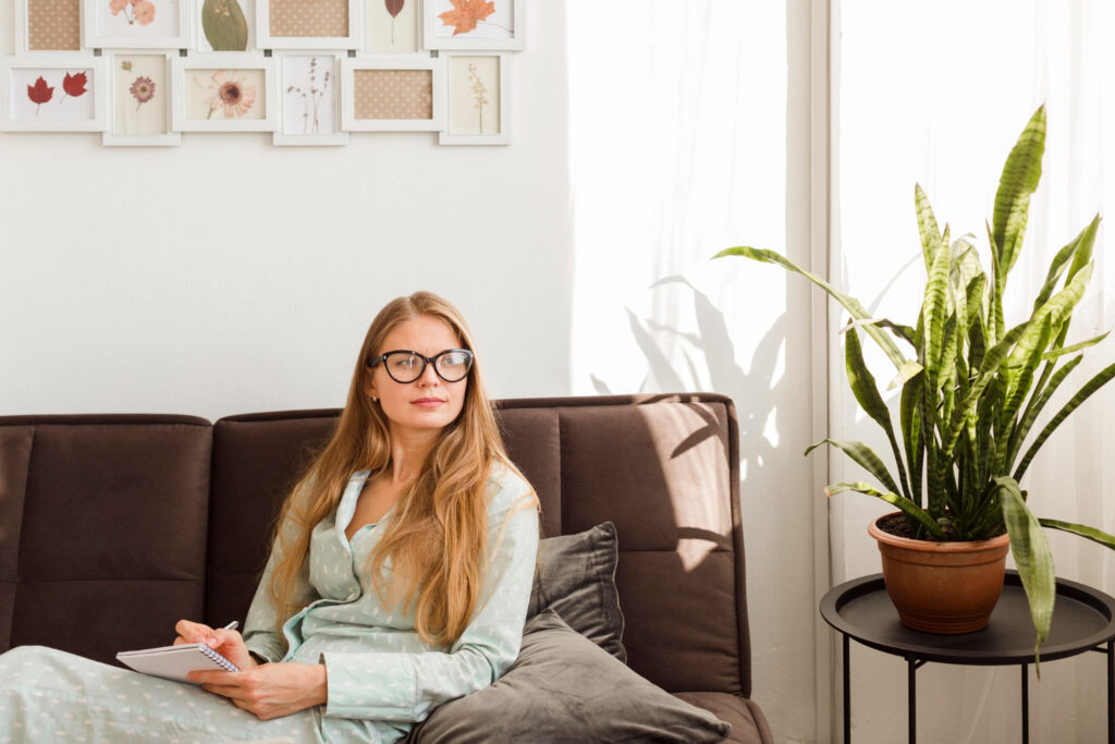 Woman in glasses, contemplating heating and cooling Guelph options, and a casual dress sitting thoughtfully on a brown couch with a notepad, in a well-lit living room with wall art, a side table, and a potted snake plant, conveying a peaceful and creative home environment.