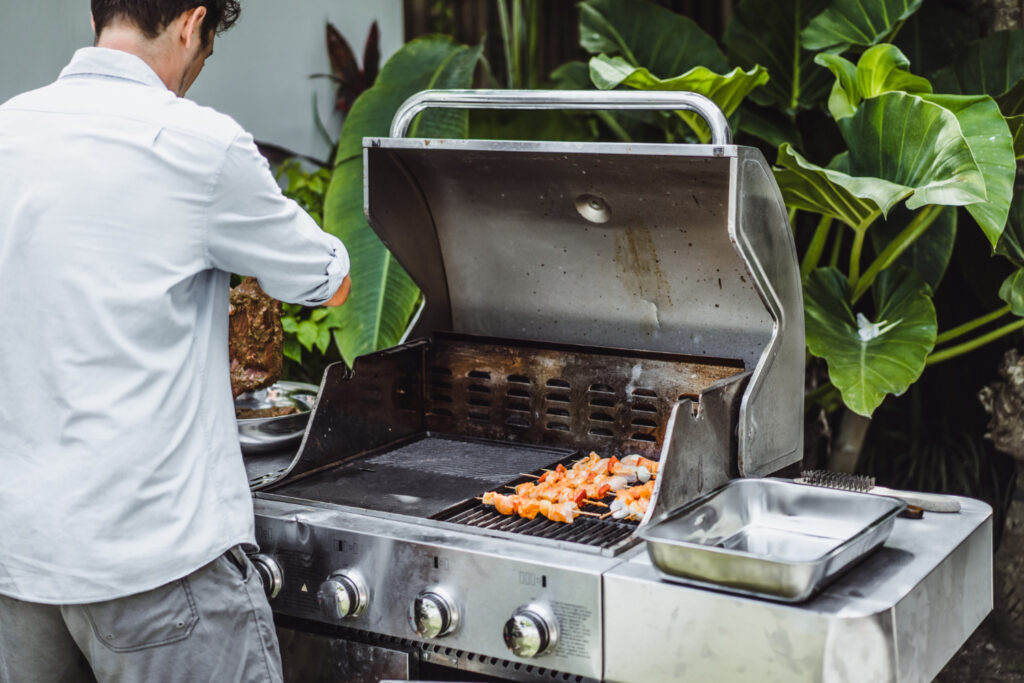 Man in a white shirt grilling shrimp skewers on a stainless steel barbecue grill, with a lush green garden in the background, after having his bbq gas line installed. 