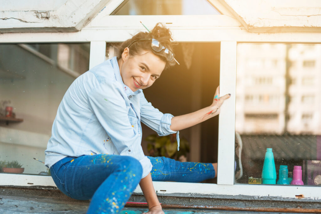 Cheerful young woman in a blue shirt and speckled blue jeans renovating an old white window frame with window caulking exterior, sunlight enhancing the casual, do-it-yourself atmosphere. Paint splatters are visible on her clothes, indicating a creative or refurbishing project in progress. Her smile and casual posture add a personal, engaging touch to the home improvement theme.