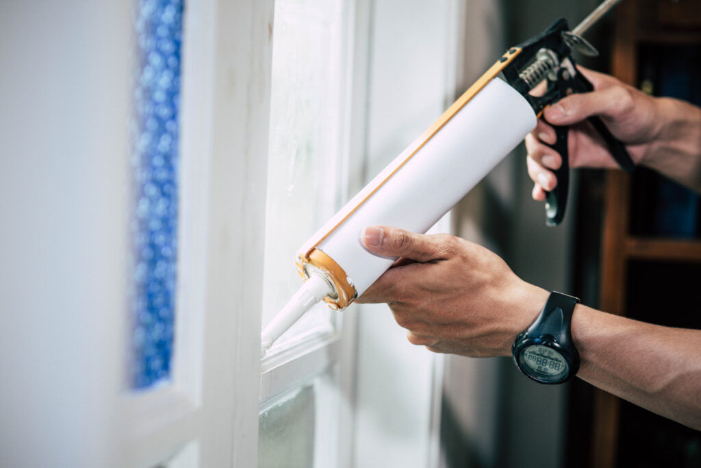 Professional hand applying caulk to seal a white window frame to improve home energy efficiency. The image shows a close-up of a person's hands using a caulking gun along the edge of a window frame, a common home improvement technique for insulation